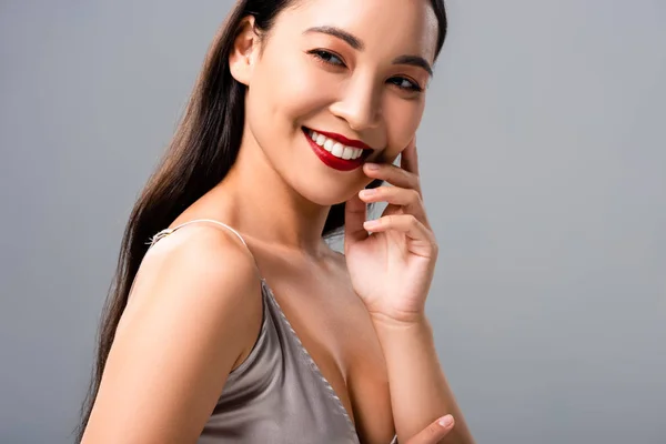 Mujer asiática feliz en vestido de satén con labios rojos aislados en gris - foto de stock