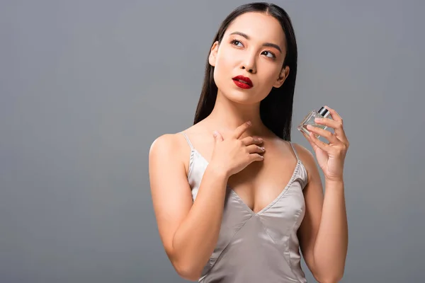 Femme asiatique rêveuse en robe élégante avec lèvres rouges tenant parfum isolé sur gris — Photo de stock