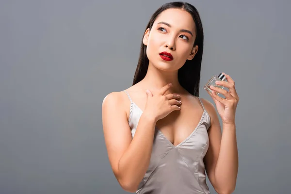 Mujer asiática soñadora en elegante vestido con labios rojos sosteniendo perfume aislado en gris - foto de stock