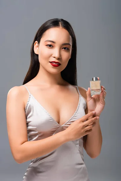 Hermosa mujer asiática en elegante vestido con labios rojos celebración de perfume aislado en gris — Stock Photo
