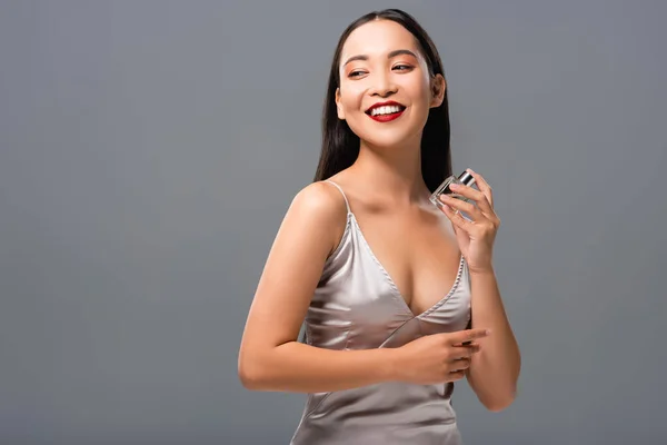 Hermosa sonriente asiático mujer con rojo labios celebración perfume aislado en gris - foto de stock