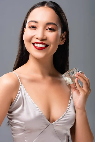 Красива усміхнена азіатська жінка в елегантній сукні з червоними губами тримає парфуми ізольовані на сірому — стокове фото