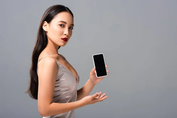 Красива азіатська жінка в атласному платті з червоними губами тримає смартфон з порожнім екраном ізольовано на сірому — стокове фото