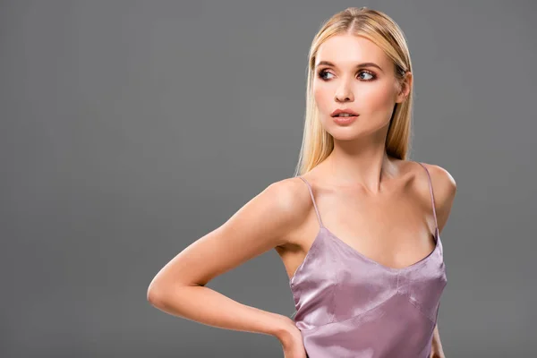 Elegante chica rubia en vestido de satén violeta con la mano en la cadera mirando hacia otro lado aislado en gris - foto de stock