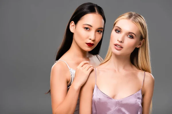 Schöne europäische blonde und asiatische brünette Frauen in Satinkleidern isoliert auf grau — Stockfoto