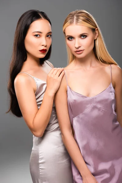 Elegant european blonde and asian brunette women in satin dresses isolated on grey — Stock Photo