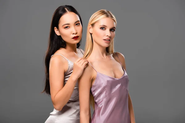 Elegante blonde und brünette Frauen in Satinkleidern schauen vereinzelt in die Kamera auf grau — Stockfoto