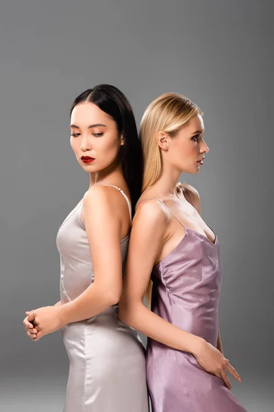 Elegante rubia y morena mujeres en vestidos de satén de pie espalda con espalda aislado en gris - foto de stock