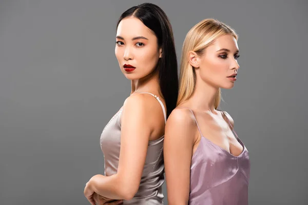 Beautiful blonde and brunette women in satin dresses standing back to back isolated on grey — Stock Photo