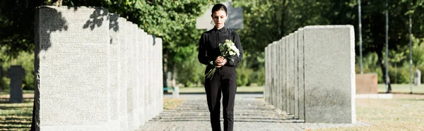 Plan panoramique de femme bouleversée tenant des fleurs et marchant dans le cimetière — Photo de stock