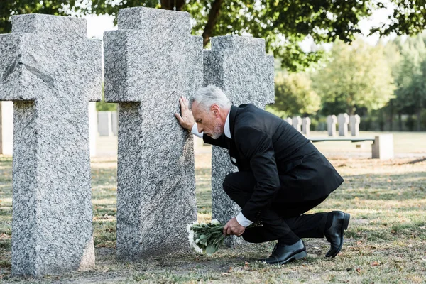 Turbato uomo anziano in giacca e cravatta seduto vicino a lapidi e in possesso di fiori — Foto stock