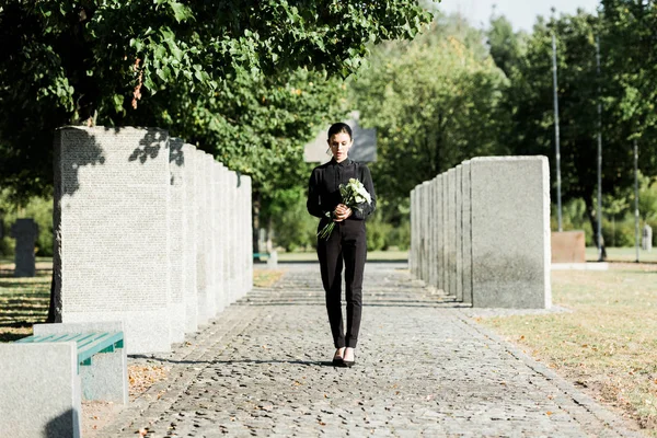 Mujer molesta sosteniendo flores y caminando en el cementerio - foto de stock