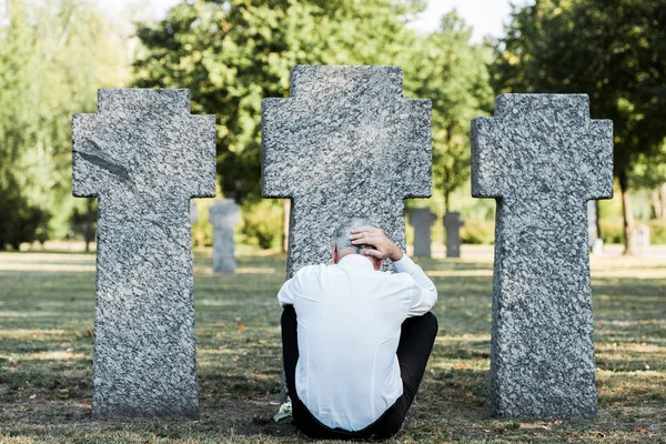 Vista trasera del hombre con el pelo gris sentado cerca de lápidas - foto de stock