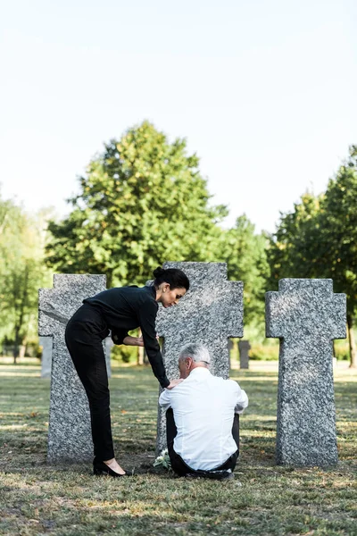 Attraente donna in piedi vicino sconvolto anziano uomo seduto vicino tombe — Foto stock