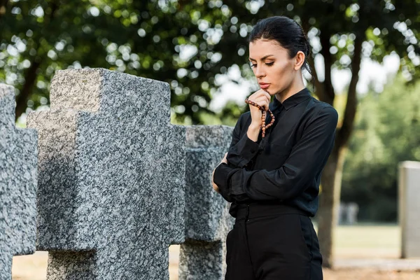 Attractive and sad woman holding rosary beads near tomb — Stock Photo