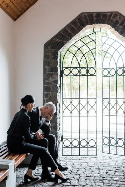 Belle femme assise sur banc près bouleversé senior homme dans cimetière — Photo de stock