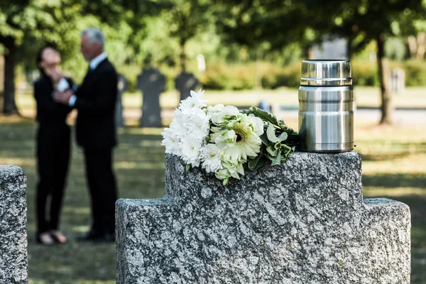 Selective focus of bouquet of white flowers and mortuary urn on tombstone near man and woman — Stock Photo