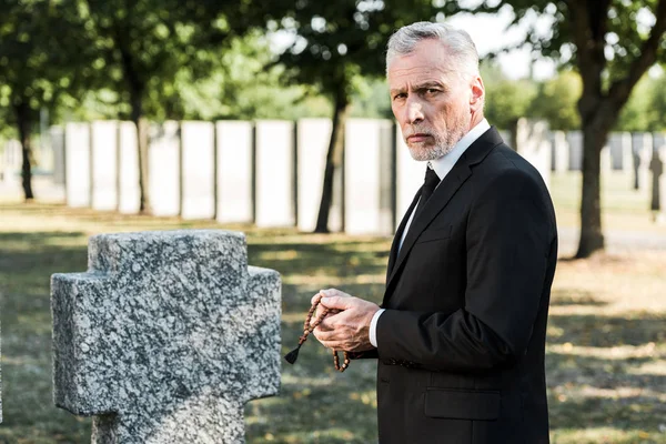 Homme avec les cheveux gris tenant chapelet perles près de la pierre tombale — Photo de stock