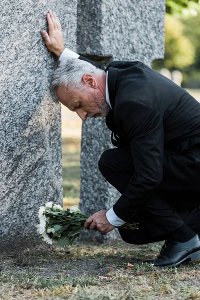 Грустный мужчина с седыми волосами кладет цветы на кладбище — стоковое фото