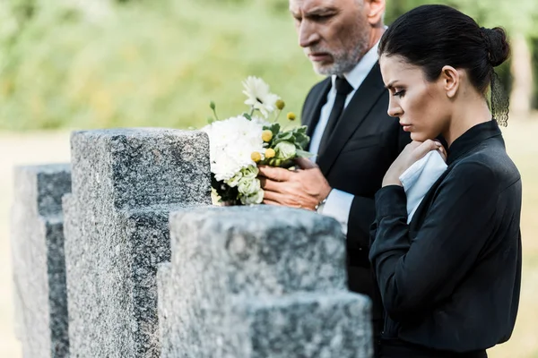 Селективное внимание мужчины с седыми волосами, держащего цветы возле надгробий и женщины — стоковое фото