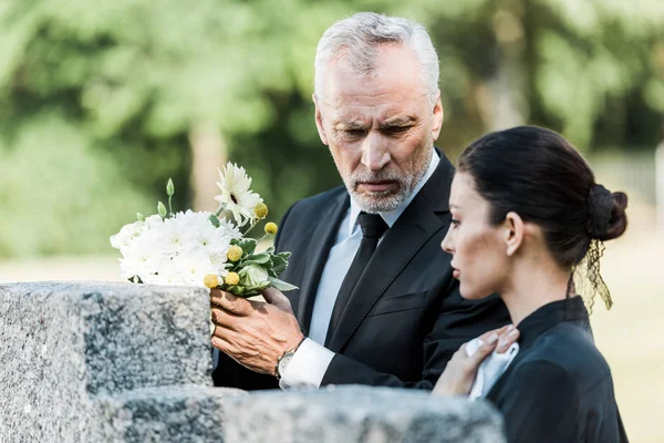 Селективное внимание расстроенного мужчины, держащего цветы и стоящего рядом с женщиной на кладбище — стоковое фото