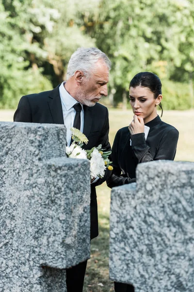 Foco seletivo de homem chateado segurando flores e olhando para a mulher pensativa no cemitério — Fotografia de Stock