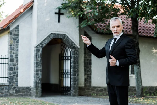 Senior man showing thumb up and pointing with finger at church — Stock Photo