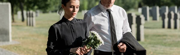 Panoramic shot of bearded senior man near woman with flowers on funeral — Stock Photo