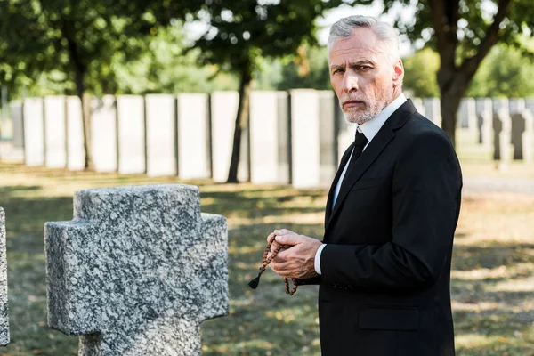 Расстроен старший мужчина держа четки возле надгробия — стоковое фото