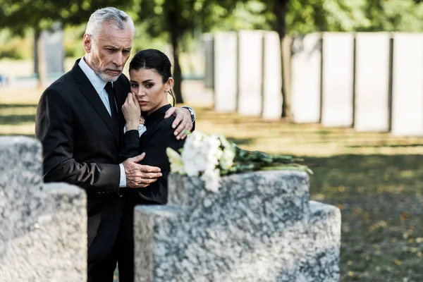 Selektiver Fokus des Mannes, der Frau umarmt, während er Blumen am Grab betrachtet — Stockfoto