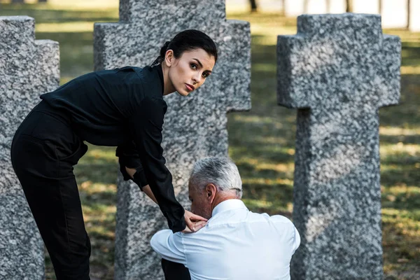 Mujer atractiva mirando a la cámara cerca de hombre triste en el cementerio - foto de stock