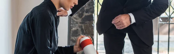 Panoramic shot of senior man standing near upset woman sitting on bench and holding mortuary urn — Stock Photo