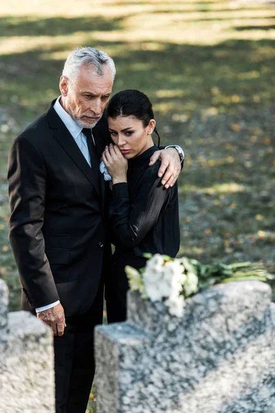 Избирательный фокус пожилого мужчины обнимающего расстроенную женщину на кладбище — стоковое фото