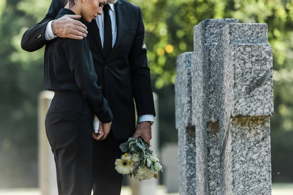 Обрезанный вид пожилого мужчины, держащего цветы, обнимающего женщину возле могил — стоковое фото