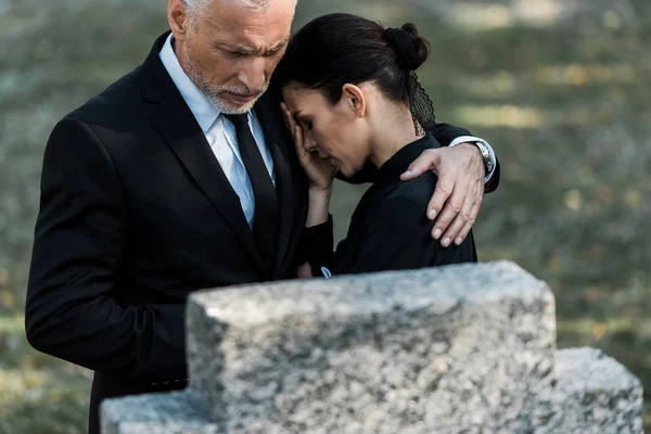 Избирательный фокус пожилого мужчины обнимающего расстроенную женщину возле надгробия — стоковое фото
