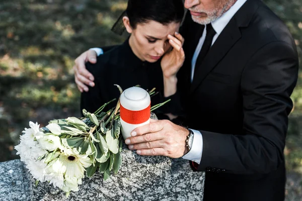 Selektiver Fokus von Blumen auf Grab in der Nähe eines älteren Mannes, der eine verärgerte Frau umarmt — Stockfoto