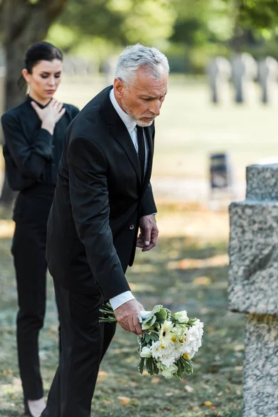 Selective focus of senior man putting flowers near tomb and woman — Stock Photo