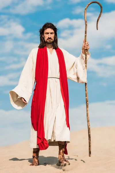 Hombre guapo en jesus bata sosteniendo bastón de madera y caminando en el desierto - foto de stock