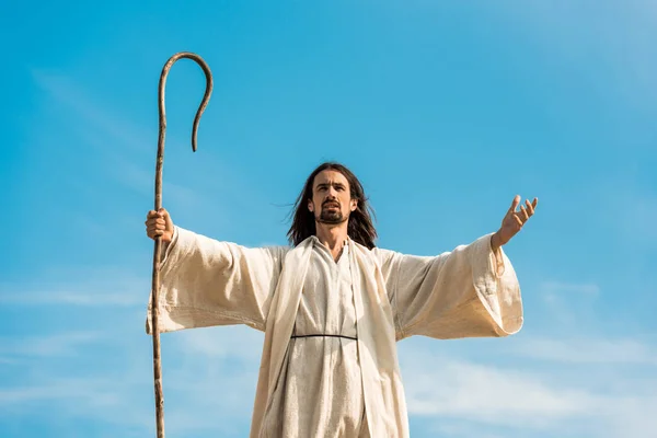 Jesús con las manos extendidas sosteniendo bastón de madera contra el cielo azul - foto de stock