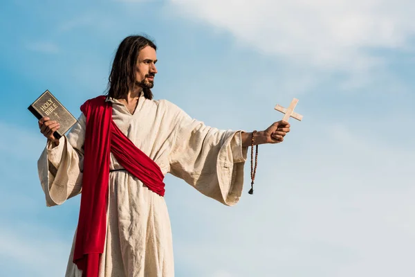 Jesus mit heiliger Bibel und Kreuz gegen blauen Himmel mit Wolken — Stockfoto