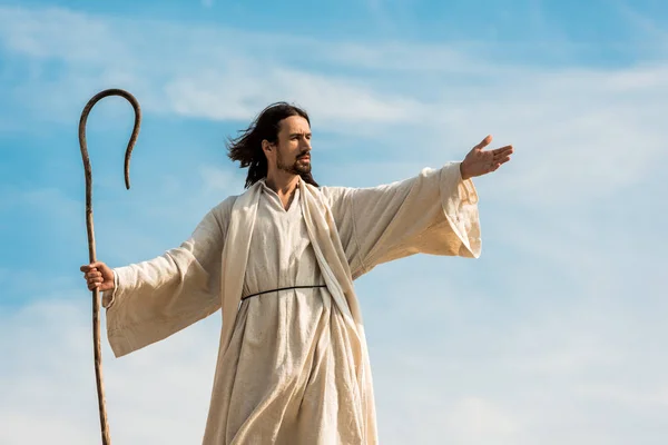 Jesús con la mano extendida sosteniendo bastón de madera contra el cielo azul - foto de stock
