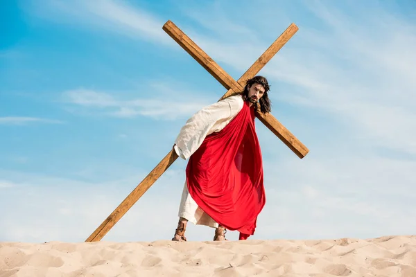 Jesús caminando con cruz de madera en el desierto contra el cielo - foto de stock