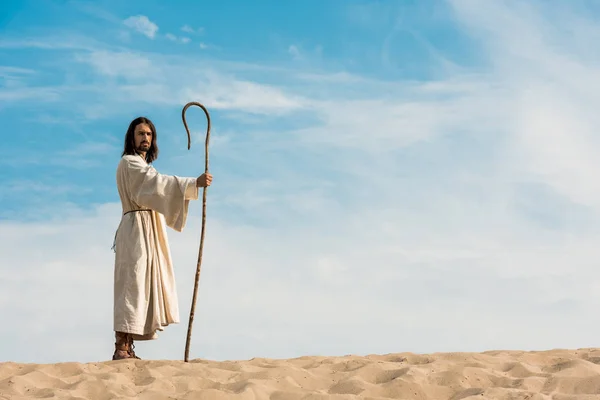 Handsome man in jesus robe holding wooden cane against sky in desert — Stock Photo