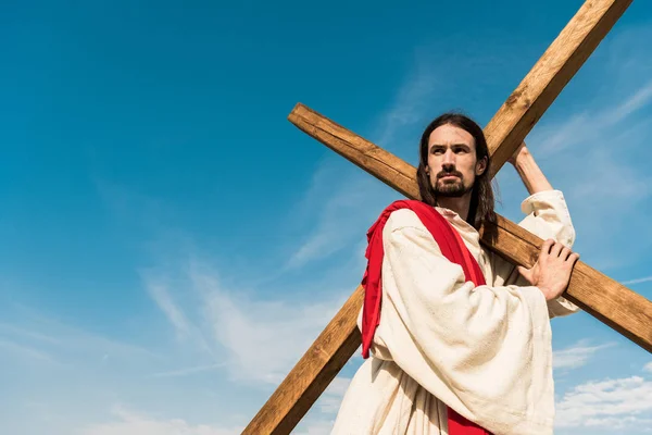 Vista basso angolo di Gesù barbuto tenendo croce contro il cielo con le nuvole — Foto stock