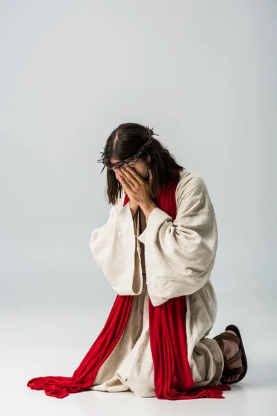 Ісус у вінку вкриває обличчя під час молитви на колінах на сірому — стокове фото