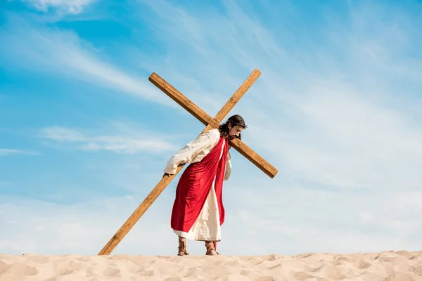 Bärtiger Mann im Jesusgewand läuft mit Holzkreuz gegen den Himmel in der Wüste — Stockfoto