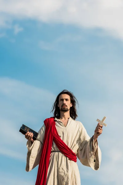 Gesù che tiene la santa bibbia e croce contro il cielo blu con le nuvole — Foto stock