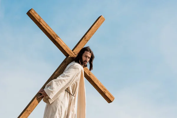 Bearded man walking with wooden cross against sky and clouds — Stock Photo