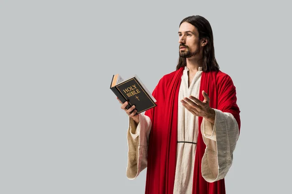 Schöner Mann gestikuliert, während er die heilige Bibel isoliert auf grau hält — Stockfoto