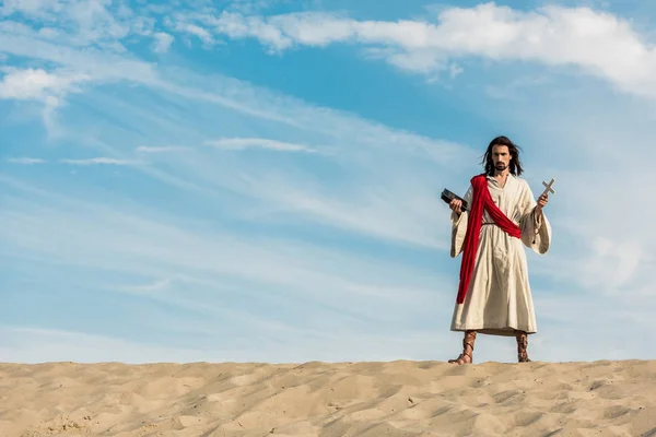 Jesus mit heiliger Bibel und Kreuz vor blauem Himmel mit Wolken in der Wüste — Stockfoto